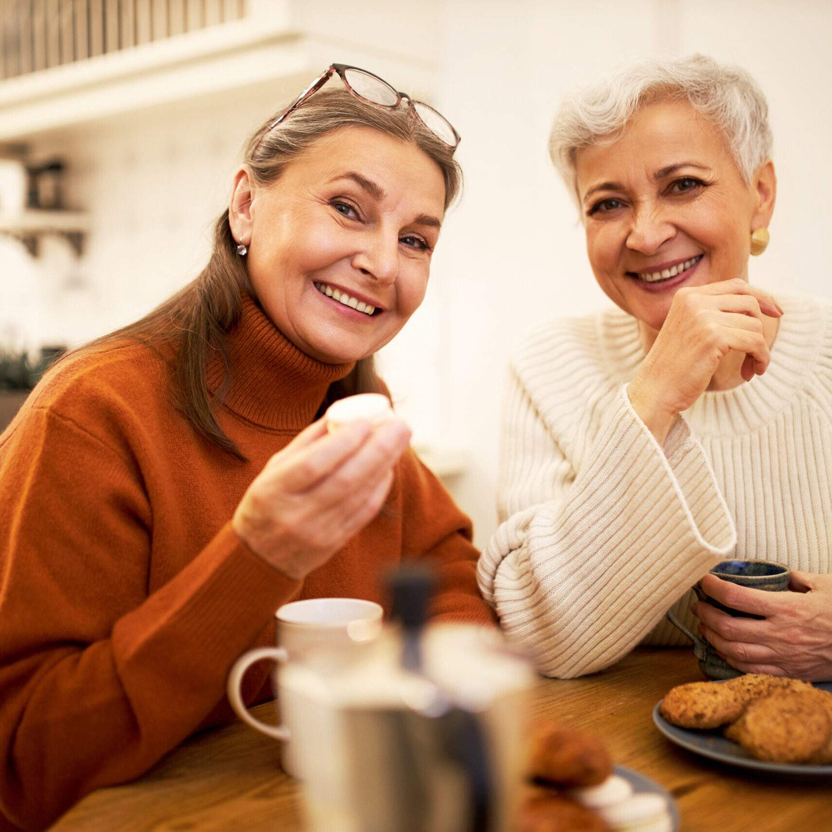 People, friendship, age and retirement concept. Indoor shot of two elegant happy women pensioners sitting at cafe table, drinking coffee and eating cookies during meeting, having nice conversation